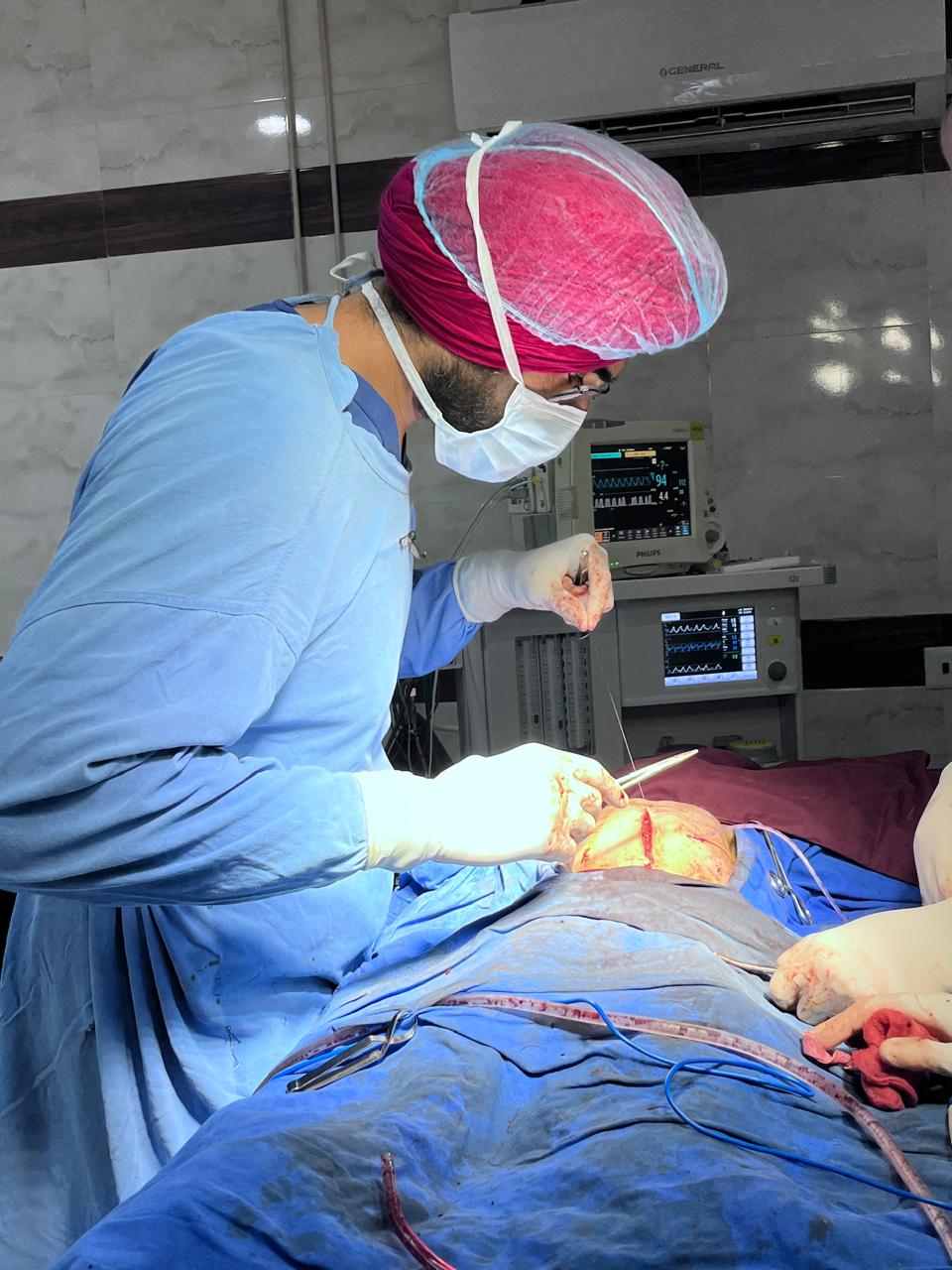 Dr. Ravitej Singh Bal suturing during a neonatal procedure, with anaesthesia monitors behind him